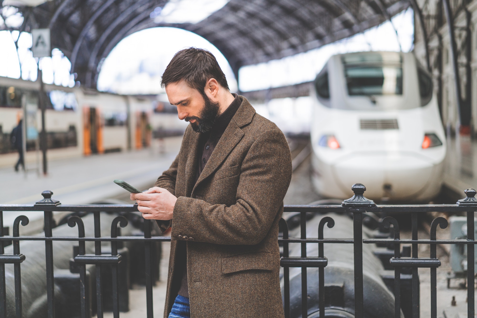 Attractive bearded man using a smart phone in his hand is standing with travel bag at the platform