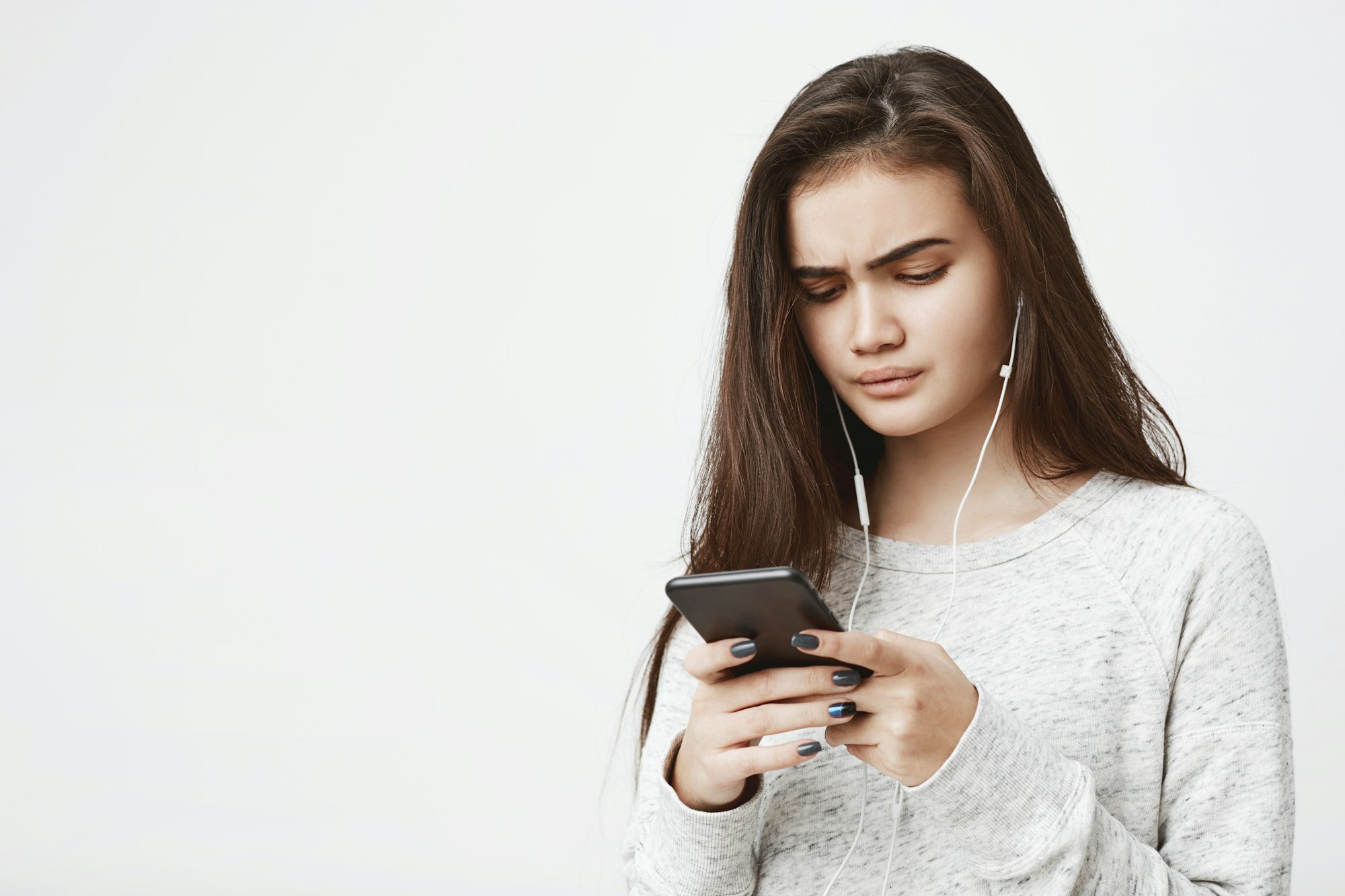 Attractive young female with worried expression holds phone and listen music, over white background
