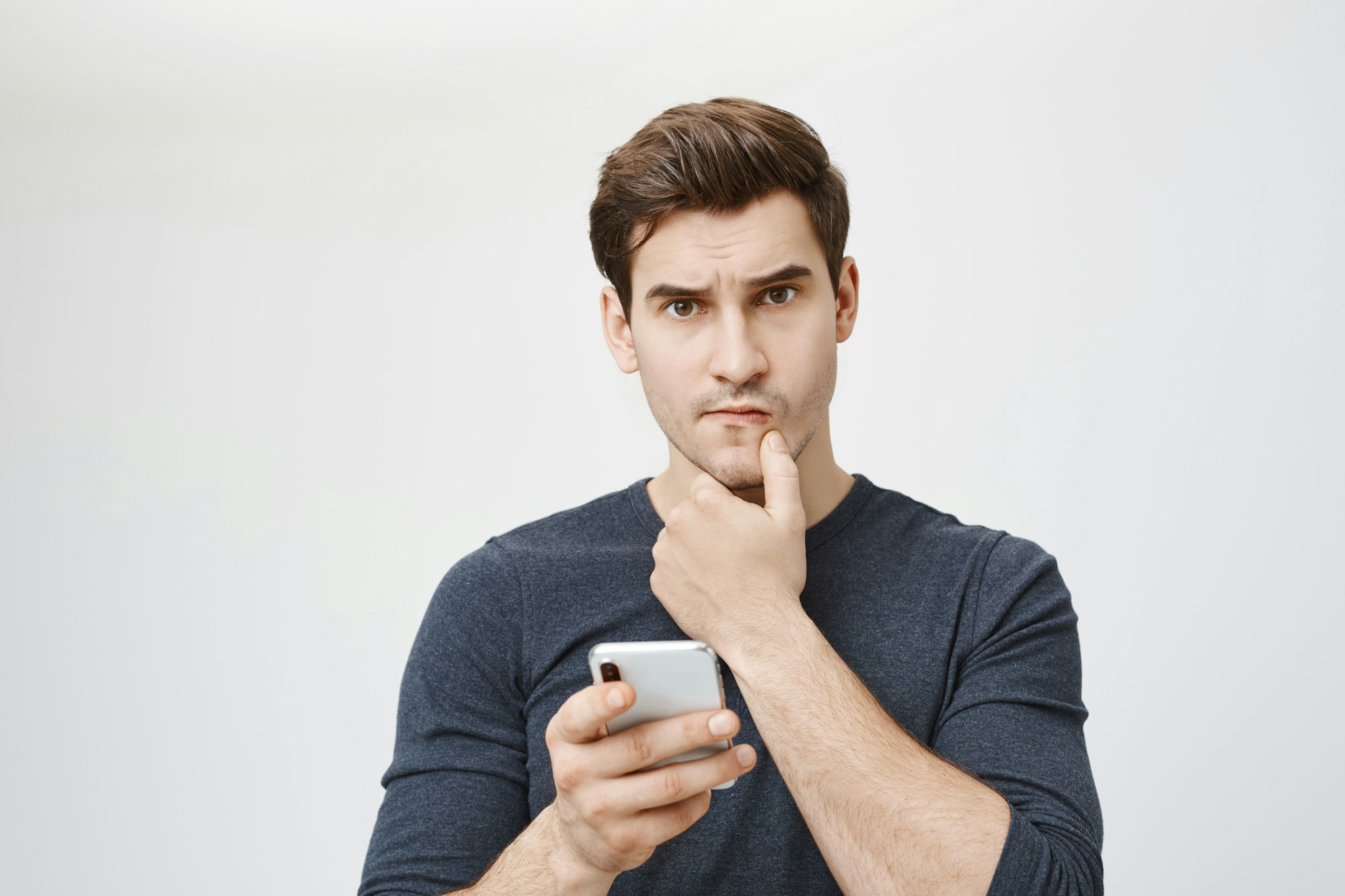 Indoor shot of young caucasian student looking worried and perplexed while holding smartphone and