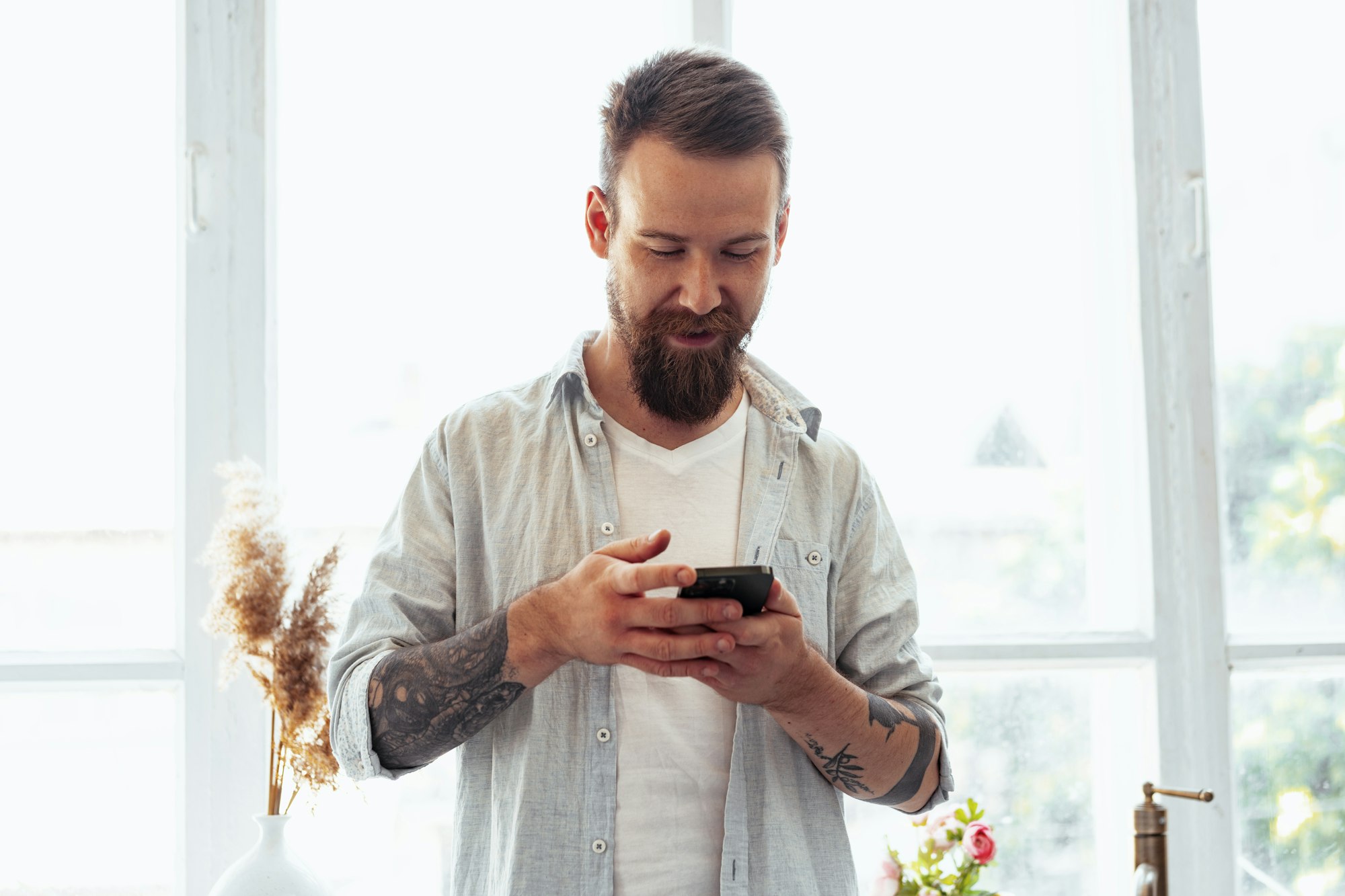 Handsome young bearded man spending time at home with phone in hands.