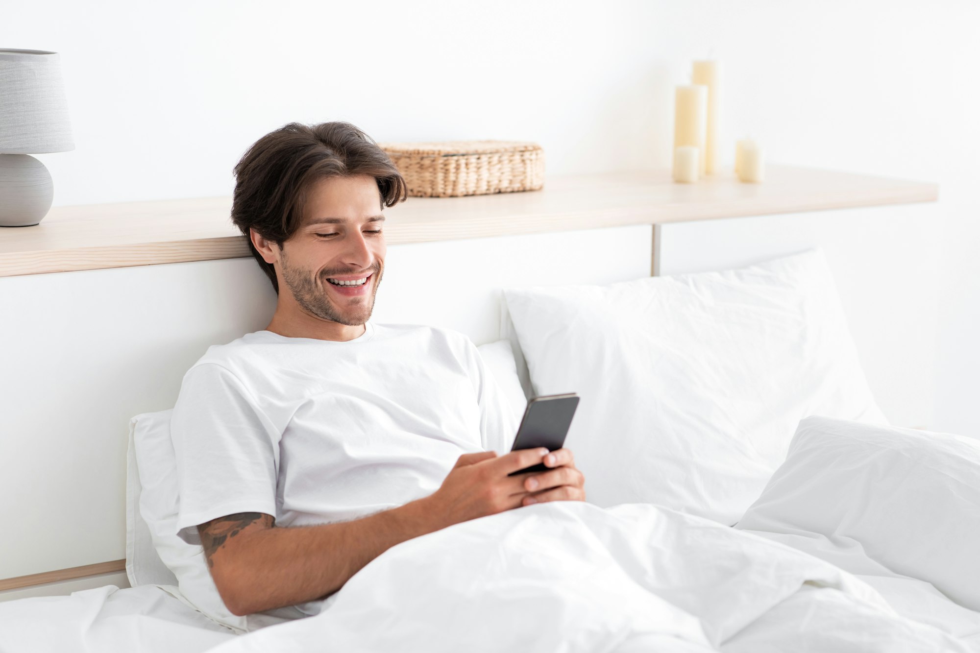 Happy european millennial man relax, typing on phone, sitting in bed in white bedroom